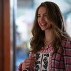 Eine lächelnde Frau in einem pink karierten Outfit und Perlenkette. Sie hält eine Tasche und wirkt glücklich.