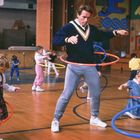In einer Turnhalle übt ein Mann mit Kindern Hula-Hoop. Basketballkorb und bunte Wandmalerei im Hintergrund.