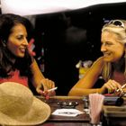 Zwei Frauen sitzen an einer Theke, lachen und halten Zigaretten. Vor ihnen liegt ein Strohhut und ein Aschenbecher.