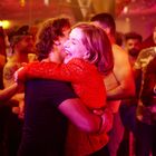 Ein festlicher, bunt beleuchteter Raum; ein lachendes Paar umarmt sich freudig inmitten einer feiernden Menge.