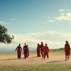 Keine erkennbaren Texte. Frauen in bunten Kleidern spazieren auf einem sandigen Platz, ein Baum links, blauer Himmel.