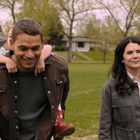 Ein Mann trägt ein Kind auf dem Rücken, während eine Frau daneben lächelt. Sie stehen auf einer grünen Wiese.