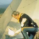 Ein Mann in Uniform kniet mit einem weißen Helm in der Hand vor einem Zelt in einer Graslandschaft.