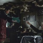 Ein Mann in rotem Overall reicht einem sitzenden Mann grüne Ofenhandschuhe, in einem Raum mit Holzwürfel-Decke.