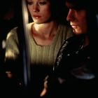 Zwei Personen sitzen nebeneinander; die linke Person schaut nach unten, die rechte hat einen ernsten Gesichtsausdruck.