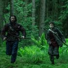 Zwei Personen mit Bögen laufen durch einen dichten, grünen Wald. Einer ist erwachsen, der andere jugendlich.