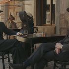 Zwei Männer in Anzügen sitzen draußen in einem Café, trinken und unterhalten sich. Retro-Kleidung und Atmosphäre.