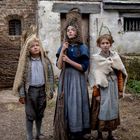 Drei Kinder in historischer Kleidung vor einer alten, rustikalen Hütte. Sie tragen ungewöhnliche Kopfbedeckungen.