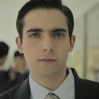 Ein junger Mann in formeller Kleidung schaut ernst in die Kamera. Hintergrund zeigt verschwommene Personengruppen.