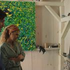 Zwei Personen betrachten bunte, abstrakte Gemälde in einem Atelier. Regale mit Malutensilien sind im Hintergrund sichtbar.