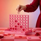 Ein rot-pinkes Spielbrett mit Herzformen und Chips, eine Hand legt einen Chip ein. Hintergrund mit warmen Tönen.