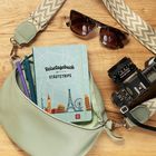 Notizbuch "Reisetagebuch Städtetrips" in Tasche mit Stiften. Daneben Sonnenbrille und Kamera auf Holzoberfläche.