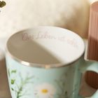"Das Leben ist schön" steht im Inneren einer Tasse, außen blumiges Design. Rechts ein vertikaler Gegenstand.