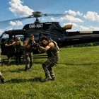 Der Text "OM-HAT AIRBUS" ist auf einem schwarzen Helikopter zu sehen. Im Vordergrund sind bewaffnete Personen in Tarnkleidung.