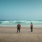 Zwei Personen mit Wanderrucksäcken stehen am Strand, blicken zum Meer mit sanften Wellen unter klarem, blauem Himmel.