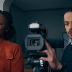 Eine Frau in orangefarbener Jacke sitzt neben einem Mann, der eine Kamera bedient und Kopfhörer trägt.