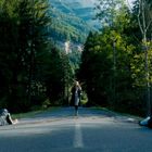 Keine Texte sichtbar. Frau geht auf leerer Landstraße auf Wald zu, zwei Personen liegen seitlich auf der Straße.