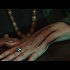 Ein Arm mit drei blutigen Kratzern; eine Hand trägt einen Ring, im Hintergrund eine Kette aus großen Perlen.