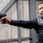 Ein Mann in schwarzer Weste hält eine Pistole mit ausgestrecktem Arm. Hintergrund: Metallstruktur.
