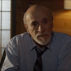 Ein älterer Mann mit Krawatte, in einem Raum mit warmem Licht und einer Lampe im Hintergrund, schaut nachdenklich.