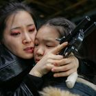 Eine Frau hält schützend ein Kind, beide wirken emotional und halten eine Waffe.