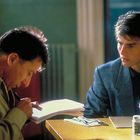 Zwei Männer sitzen an einem Tisch. Einer liest in einem Notizbuch; auf dem Tisch stehen Gewürzbehälter.