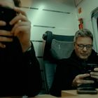 Zwei Männer in einem Zugabteil, beide schauen auf ihre Smartphones. Einer sitzt auf einem Platz mit der Nummer 1.