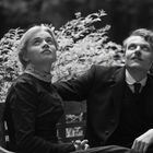 Zwei Personen in formeller Kleidung sitzen auf einer Bank und blicken nach oben, umgeben von Pflanzen.