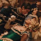 Ein Paar in historischer Kleidung tanzt elegant in einem prunkvollen Ballsaal, umgeben von weiteren Tanzpaaren.