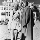 Zwei Personen in Bademänteln am Strand vor einem großen, mehrstöckigen Gebäude. Sie lächeln und stehen im Sand.