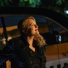 Eine Frau mit blondem Haar steht lächelnd neben einem Auto bei Nacht, im Hintergrund sind Bäume erkennbar.