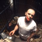 Auf einem dekorativen Boden steht ein Mann im blutbefleckten Shirt mit "B5460-8", daneben ein Tablett und eine Person liegt am Boden.