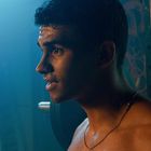 Ein verschwitzter Mann mit Kette steht in einem Raum mit Trainingsgeräten, gedämpftes Licht.