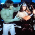 Dunkelgrüner, muskulöser Mann mit wildem Haar neben großem, rätselhaftem Krieger in Rüstung und Fell.