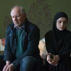 Ein Mann und eine Frau sitzen nebeneinander auf einem Bett. Der Mann schaut ernst, die Frau trägt ein schwarzes Kopftuch.