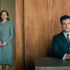 Eine Frau steht in einem grauen Kleid in einer Holztüröffnung. Ein Mann sitzt nachdenklich an einem Tisch.