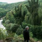 Ein Mann steht auf einem Hügel, blickt auf einen Fluss, umgeben von dichtem Wald. Eine Schattenhand erstreckt sich über die Bäume.