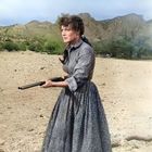 Eine Frau in einem langen Kleid steht in einer Wüstenlandschaft und hält ein Gewehr.