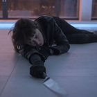 Eine Person liegt am Boden und streckt die Hand nach einem Messer aus. Sie trägt eine Lederjacke und sieht erschöpft aus.