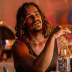 Ein Mann mit langen Haaren und Bart sitzt entspannt an einem Tisch mit Getränkeflaschen, düster beleuchtet.