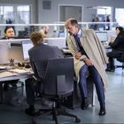 Mehrere Personen arbeiten an Schreibtischen in einem Großraumbüro. Ein Mann im Trenchcoat spricht mit einem Kollegen.