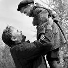 Ein lächelnder Mann hebt ein Kind hoch, beide in Winterkleidung. Der Hintergrund ist unscharf mit Bäumen.