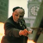 Eine Person mit einer entstellten Maske hält eine Pistole, Hintergrund zeigt eine Explosion und ein unscharfes Uhrenturm.