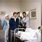 Vier Männer in bunter Kleidung betrachten jemanden in einem Krankenhausbett. An den Wänden hängen zwei Landschaftsgemälde.