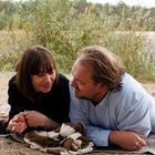 Zwei Menschen liegen entspannt auf Decken am Sandstrand, im Hintergrund sind Pflanzen und ein Gewässer sichtbar.