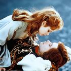 Ein Mädchen mit roten Haaren beugt sich über einen Jungen in mittelalterlicher Kleidung vor einem blauen Hintergrund.