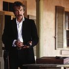 Eine Person in einem schwarzen Anzug steht auf einer Veranda, hält etwas in den Händen. Daneben ein Tisch mit einer Holzbox.