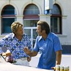 Zwei Männer in gemusterten Hemden sprechen an einem Auto. Auf dem Auto stehen Dosen mit der Aufschrift "Olympia Beer".