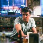 Ein Mann in einem grauen T-Shirt lehnt an einer Bar. Im Hintergrund bunte Neonlichter und Getränkekisten.