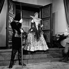 Eine Frau in einem opulenten Kleid tritt aus einer prächtigen Tür, begleitet von einer Wache mit Speer.